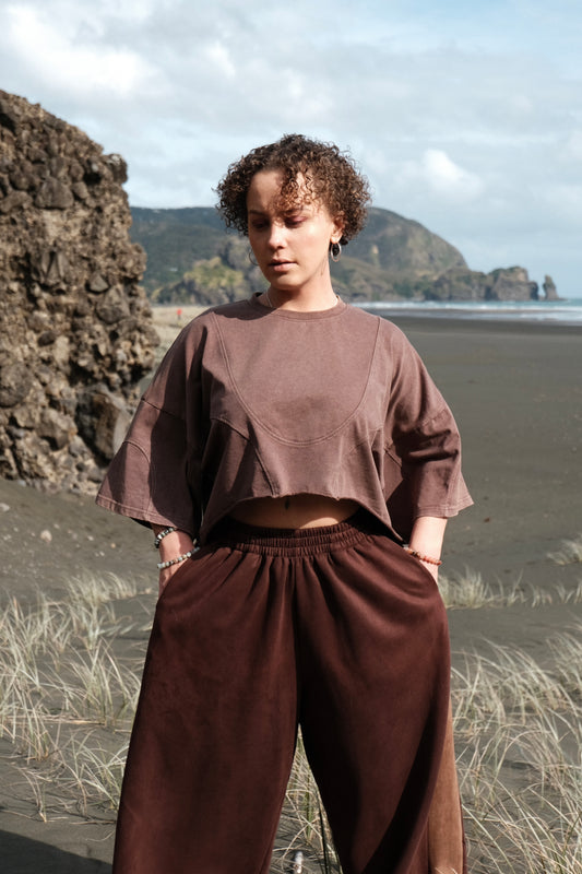 Person wearing a brown outfit standing on a beach with rocky formations and water in the background