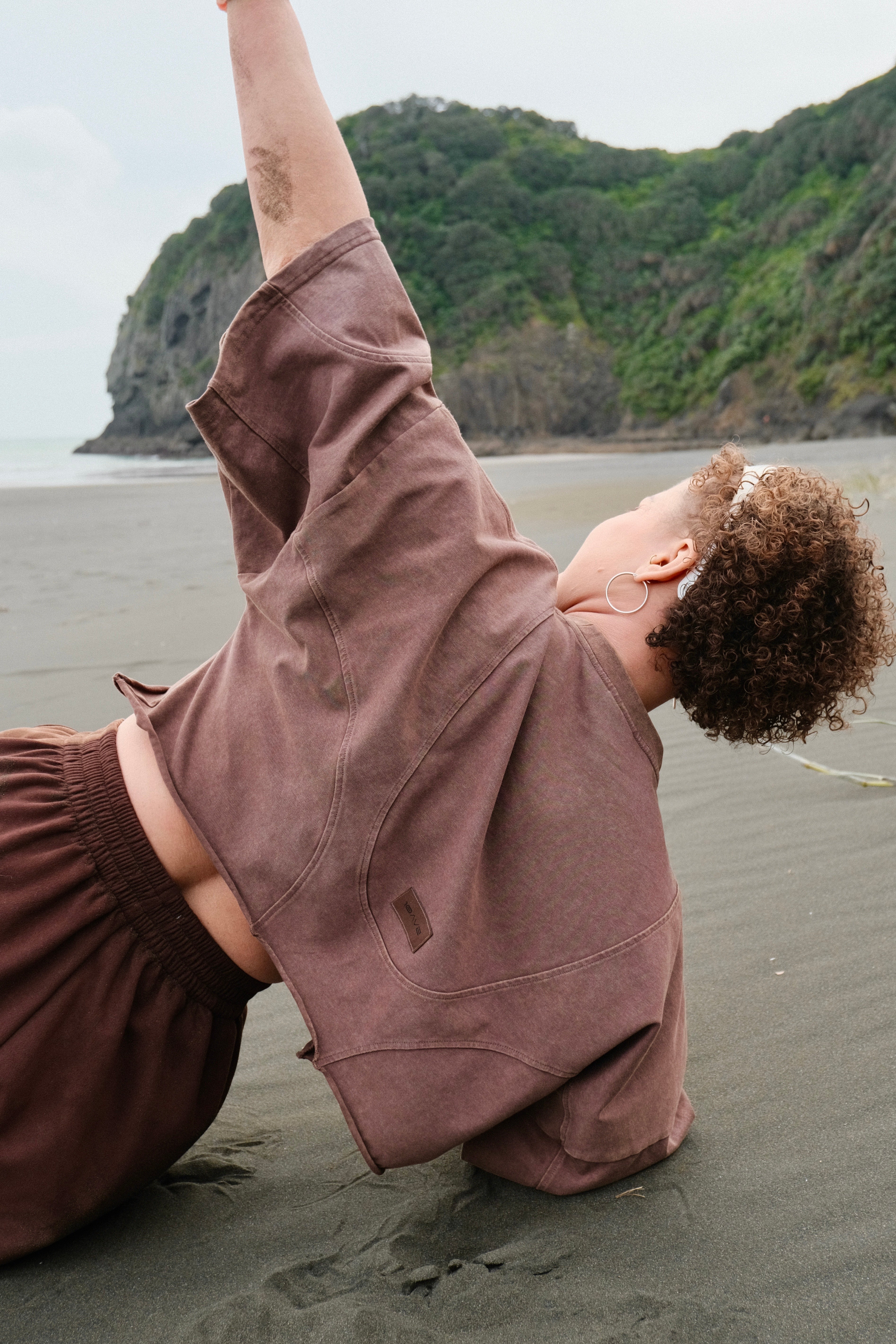 Person in a brown outfit stretching on a beach with green hills in the background