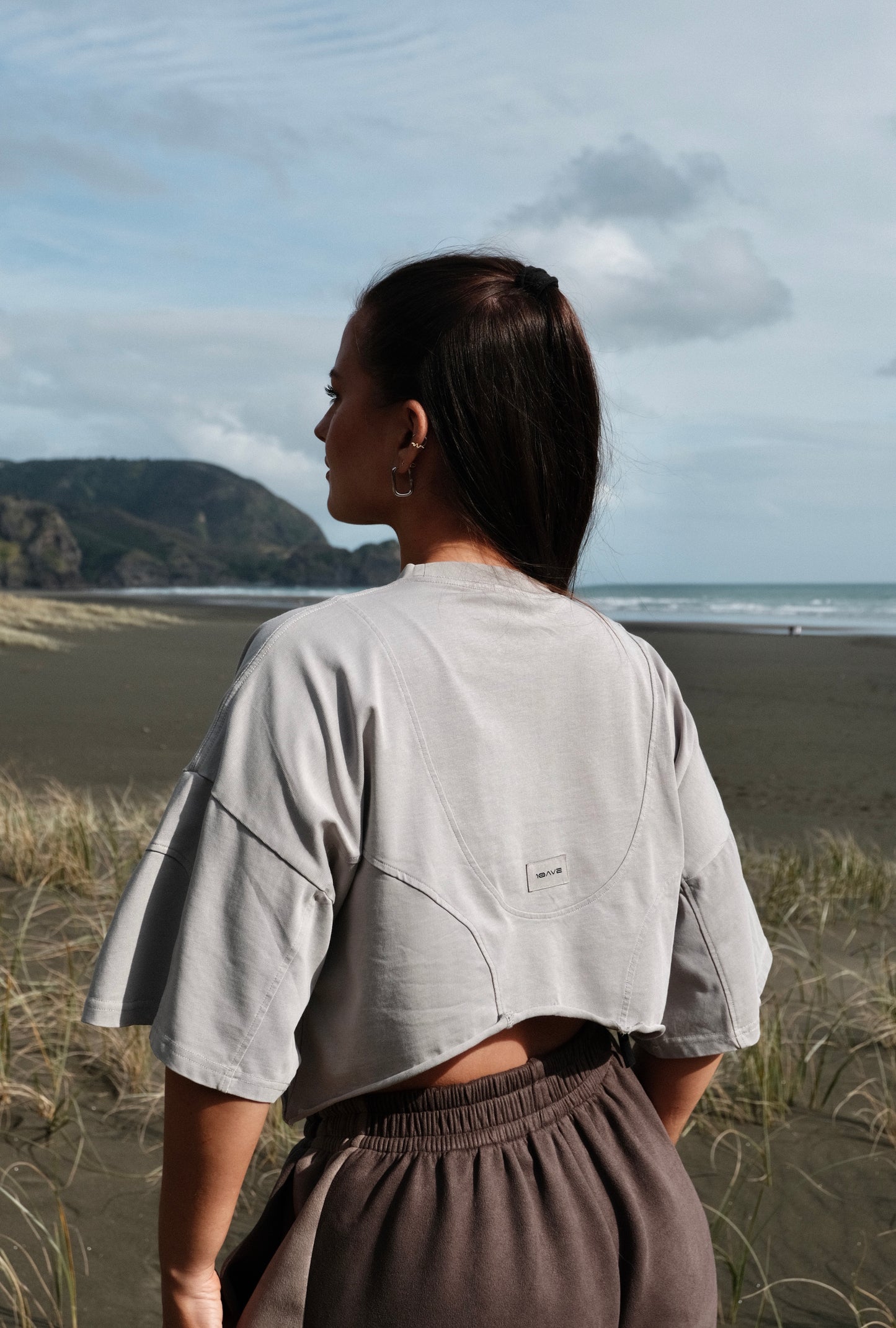 Person wearing a light gray oversized shirt and gray pants standing in a grassy field with a scenic background.
