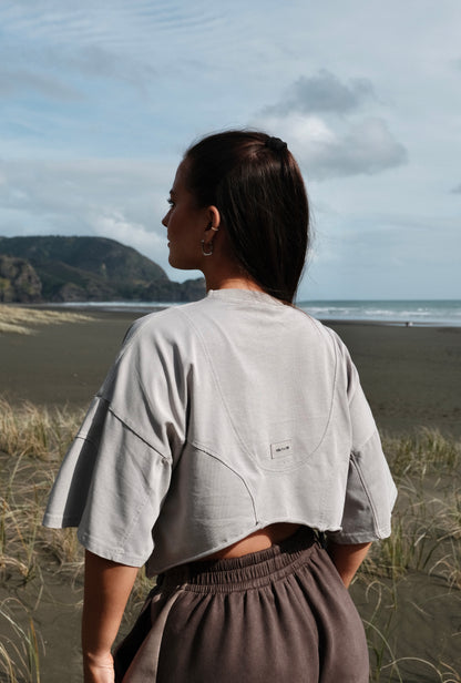 Person wearing a light gray oversized shirt and gray pants standing in a grassy field with a scenic background.