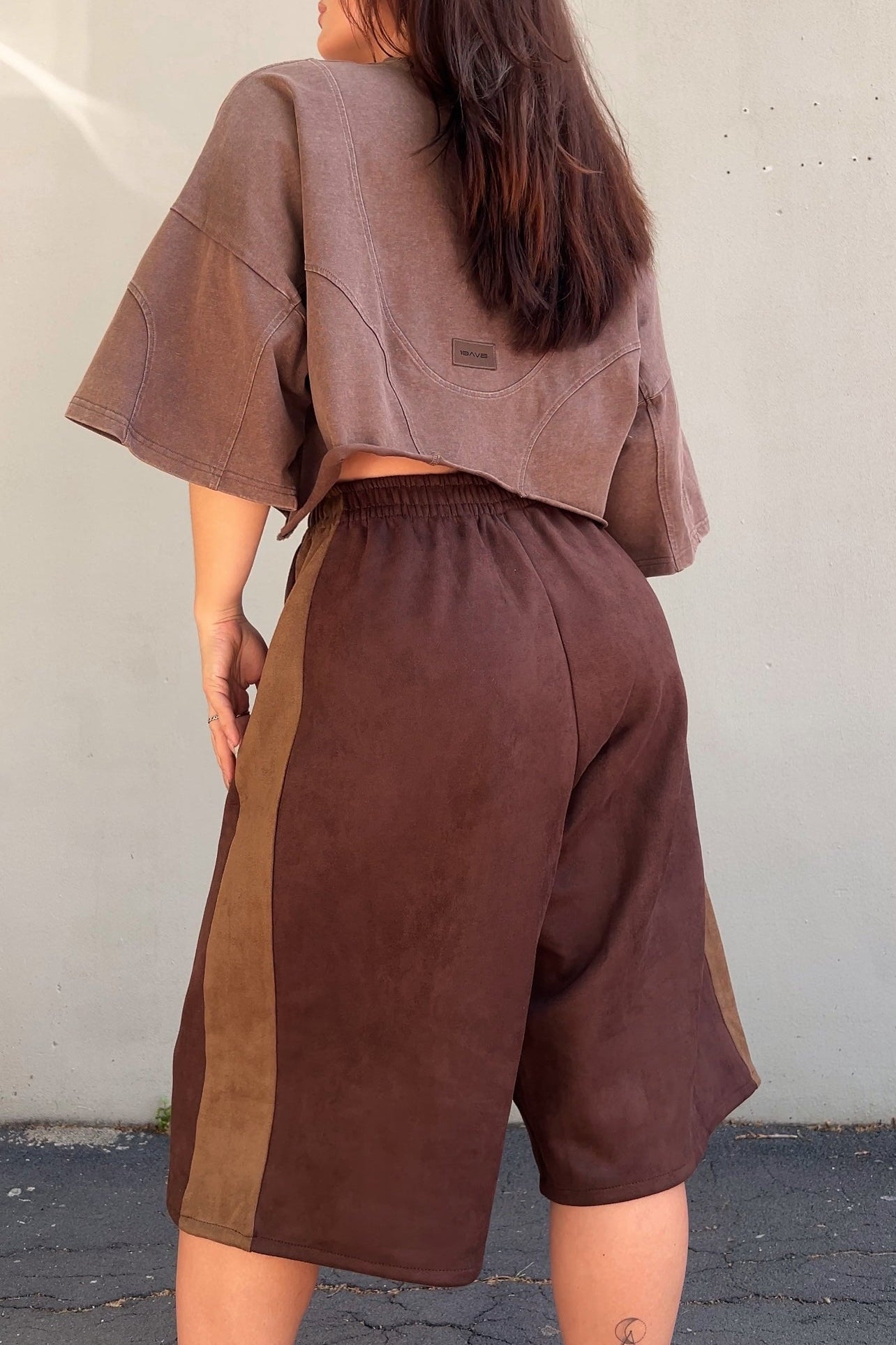 Person wearing a brown outfit with a t-shirt and jorts against a white wall.
