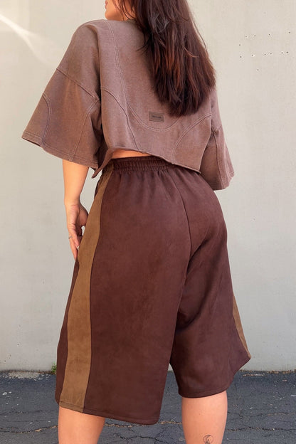 Person wearing a brown outfit with a t-shirt and jorts against a white wall.