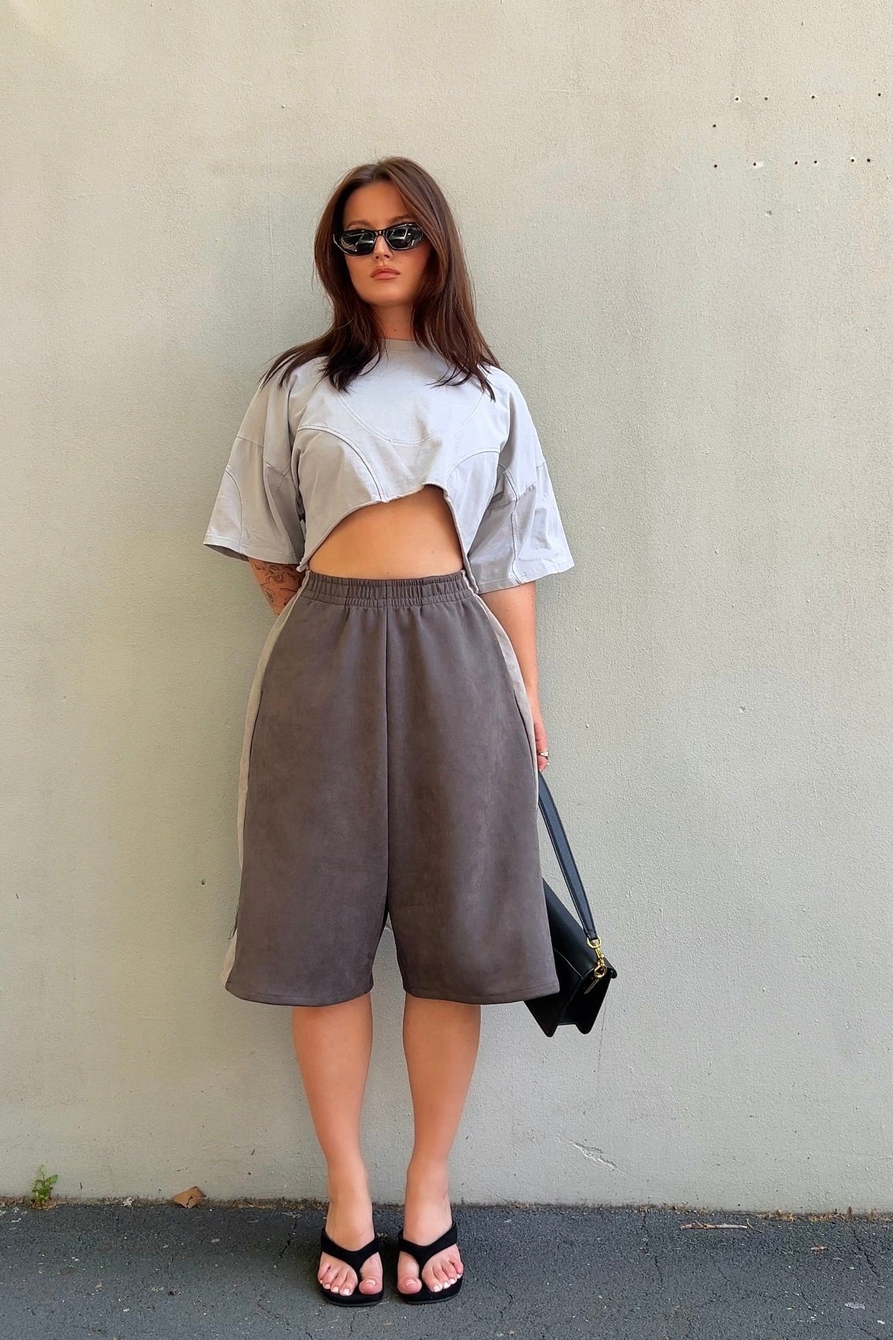 Woman wearing a white top and gray high-waisted pants against a plain wall.